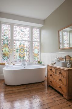 a bath tub sitting next to a large window in a bathroom with wooden floors and white walls