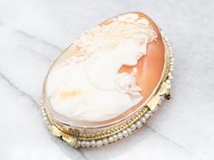 "This cameo features wonderful, crisp details, and a great contrast between the creamy white carving and the soft russet of the background. A string of seed pearls pulls the piece together, drawing on the color of the profile and the floral details of the gold. Metal: 14K Yellow Gold Gem: Shell Cameo Gem Measurements: 34.4 x 45.6 mm, Oval Accents: Seed Pearls Measurements: 40 x 55 mm Marks: \"X\" Stamped To view a video of this piece check out the link below: https://vimeo.com/802860432 SKU #: A Exquisite White Brooch For Formal Occasions, Elegant Formal Brooch With Intaglio Detail, Elegant Intaglio Brooch For Formal Occasions, White Cameo Brooch For Formal Occasions, White Cameo Brooches For Formal Occasion, Luxury Cameo Brooches, Luxury Cameo Brooch For Wedding, Luxury Intaglio Brooch For Formal Occasions, Heirloom White Brooch For Formal Occasions