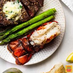 steak, lobster and asparagus on a white plate with lemon wedges next to it