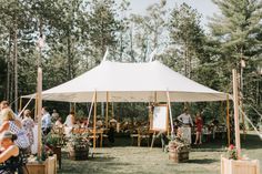 a group of people standing under a white tent in the middle of a field next to trees