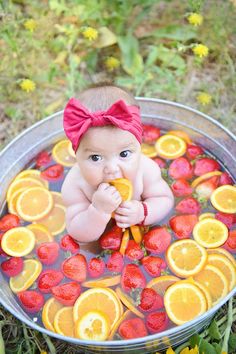 a baby sitting in a metal bowl filled with oranges and strawberries on the ground