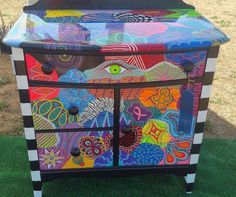 an old dresser painted with colorful designs on the top and bottom, sitting in grass