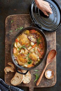 a skillet filled with chicken and vegetables on top of a cutting board next to bread