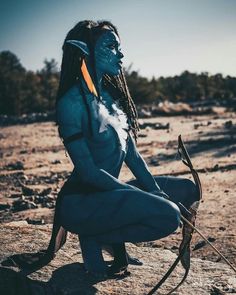 a woman with blue paint on her face and body sitting in the desert holding a bow