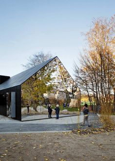 two people standing in front of a black building with trees around it and one person walking on the sidewalk