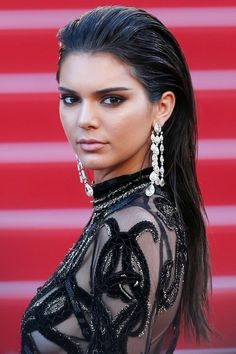 a woman with long brown hair wearing black dress and dangling earrings on her head, standing in front of a red carpet