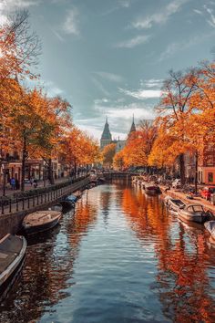 boats are parked along the side of a canal with autumn trees lining both sides and a church steeple in the distance