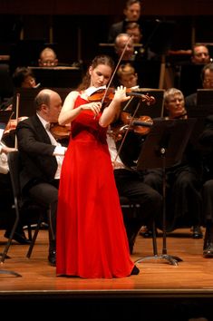 a woman in a red dress playing the violin on stage with orchestra members behind her