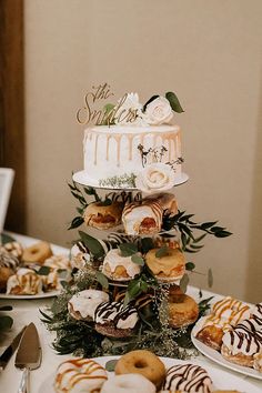 a wedding cake with donuts and greenery on the top is surrounded by other desserts