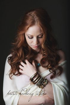 a woman with long red hair wearing gold bracelets and a white dress is looking down