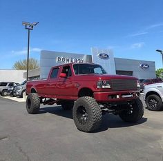 a red pickup truck parked in front of a dealership with large tires on it