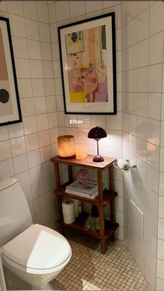 a white toilet sitting next to a wooden shelf in a bathroom under a painting on the wall