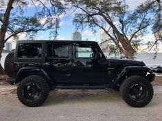 a black jeep is parked in front of some trees and the water on a sunny day