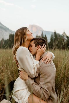 a man and woman are sitting in tall grass with mountains in the backgroud