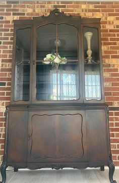 an old china cabinet with glass doors and flowers in the top shelf is shown against a brick wall