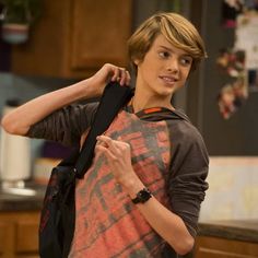 a young boy with a backpack on his back standing in a kitchen, looking at the camera