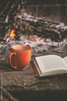 an open book sitting next to a coffee cup on top of a wooden table with a fire in the background