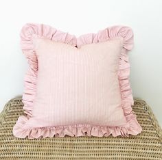 a pink pillow with ruffled edges sitting on a wicker bench next to a white wall