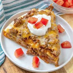 a piece of mexican casserole with sour cream on top and tomatoes in the background