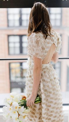 a woman in a dress holding a bouquet of flowers looking out a window at the city