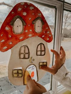 a person holding up a paper cut out of a mushroom house in front of a window