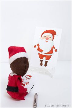 a santa clause doll sitting next to a christmas card and marker with the word santa claus on it