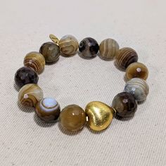 This Ro & Gieo bracelet features stunning banded agate beads paired with a chunky 24 karat gold-plated brass heart. The unique combination of materials offers a luxurious and trendy look, making it the perfect accessory for any occasion. The banded agate beads provide a natural and earthy touch, while the gold-plated heart adds a touch of elegance. Elevate your style with this exquisite bracelet. Ro & Gieo Banded Agate Bead and 24 karat gold-plated 100% brass heart chunky bracelet Beads - 16 mm Gold Agate Beaded Bracelets With Gemstones, Gold Agate Gemstone Beaded Bracelets, Gold Agate Gemstone Beaded Bracelet, Gold Agate Gemstone Bracelets, Gold Agate Bracelet With Gemstone Beads, Gold Heart-shaped Gemstone Bracelets, Gold Bracelets With Agate And Natural Stones, Gold Bohemian Bracelet With Heart Beads, Bohemian Gold Bracelets With Heart Beads