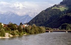 a river with mountains in the background and houses on either side, near a bridge