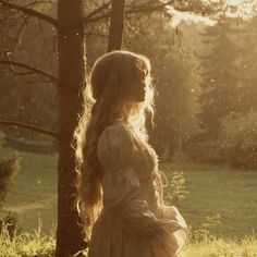 a woman with long hair standing next to a tree in a field near grass and trees