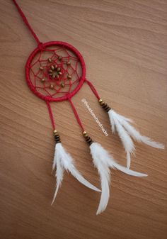 a red dream catcher with white feathers and beads on it sitting on a wooden table