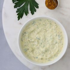 a white bowl filled with green sauce next to some parsley on a marble table