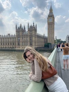 a woman leaning on the edge of a bridge next to a body of water with a clock tower in the background