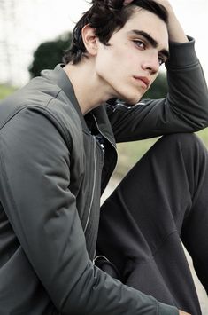 a young man sitting on the ground with his head in his hands and wearing a jacket