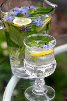 two glasses filled with water, lemon and blue flowers on a glass table outside in the sun