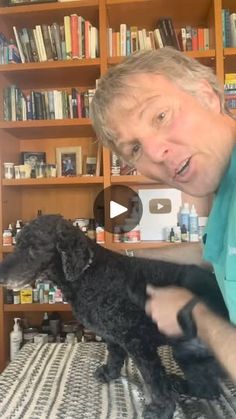 a man holding a small black dog on top of a bed in front of bookshelves