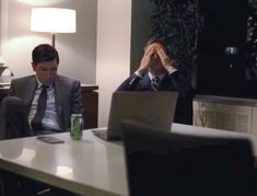 two men in suits sitting at a table with their hands to their faces and one is covering his eyes