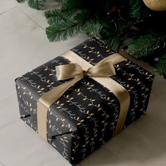 a black present wrapped in gold ribbon next to a christmas tree