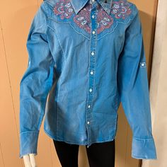 New Grace Buttoned Down Women's Blouse. Material Is Turquoise Blue With A Worn-In Look. Fabric Is Regular Weight. A Little Bling Within The Pink Embroidered Flowers. Tailored To Show Off Your Figure. Hem Length (From Collar) = 29". Wear Tucked In Or Out. Back Also Has Designed Yoke. Comes In Medium, Large And Extra Large. Western Long Sleeve Top With Floral Embroidery, Embroidered Blue Denim Blouse, Blue Embroidered Denim Blouse, Fitted Medium Wash Cotton Blouse, Blue Denim Embroidered Blouse, Fitted Cotton Blouse In Medium Wash, Blue Denim Long Sleeve Top, Fitted Denim Blue Cotton Blouse, Medium Wash Fitted Western Tops