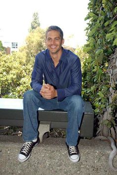 a man sitting on top of a bench in front of trees and bushes with his legs crossed