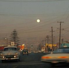 an old car driving down the road in front of other cars and power lines at dusk