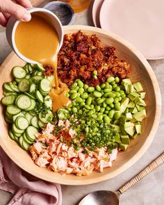 a person pouring dressing into a bowl filled with vegetables and cucumbers, on top of a table