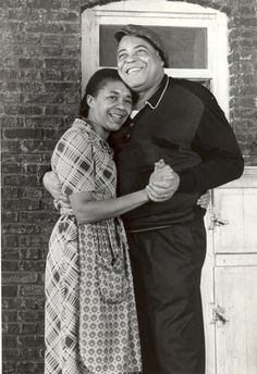 a man and woman hugging each other in front of a brick wall with an open window