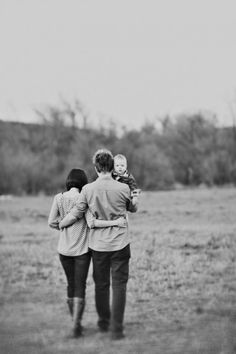 a man and woman walking through an open field with a baby in their arms while holding onto each other's shoulders