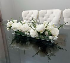 white flowers are arranged in a clear vase on a glass table with chairs behind it