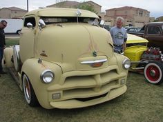 two men standing next to an old yellow truck on display at a car show with other antique cars in the background