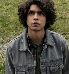 a young man with curly hair standing in front of a grassy field and looking at the camera