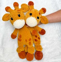 two stuffed giraffes sitting next to each other on a white sheet with one being held by someone's hand