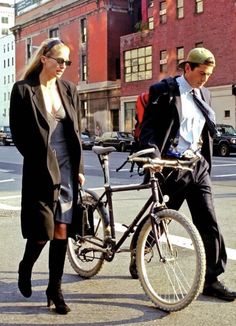 a man and woman standing next to a bike on the side of the road with buildings in the background