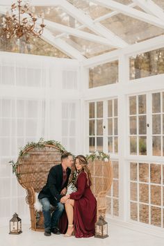 an engaged couple sitting on a wicker chair in the middle of a white room