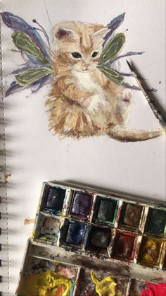 a cat sitting on top of a table next to some paint and watercolors